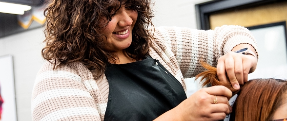 A smiling OTECH Cosmetology student styles a client's hair in a professional salon setting, demonstrating the program's real-world service experience