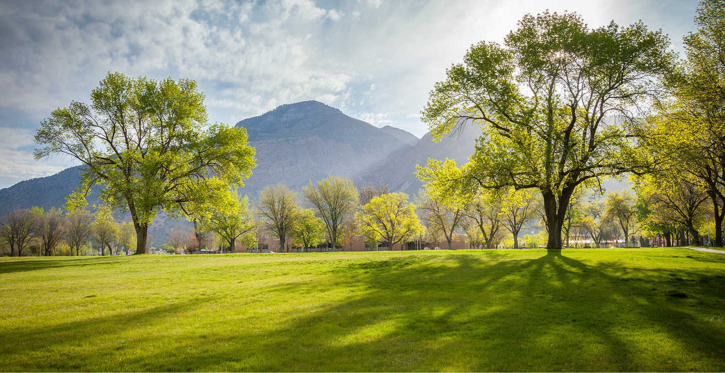 Scenic outdoor grounds of Ogden-Weber Technical College with lush green grass, large trees, and mountains visible in the background, showcasing the OTECH campus environment for community campus services
