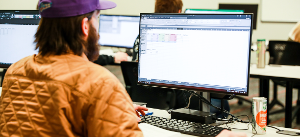 A student viewed from behind wearing a tan jacket and purple cap works at a computer workstation displaying a spreadsheet, representing the type of custom-fit workforce training OTECH provides to employer partners