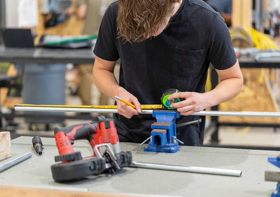 A young male apprentice measures a metal rod clamped in a workbench vise using a tape measure, with power tools including a drill nearby, representing hands-on trade skills learned through OTECH's employer-sponsored apprenticeship programs