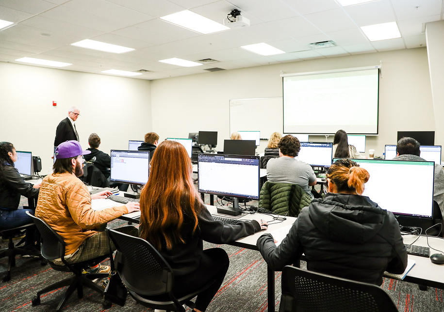 Students work at computer workstations in a training classroom at OTECH while an instructor stands at the front of the room, representing custom workforce training delivered to employer partners