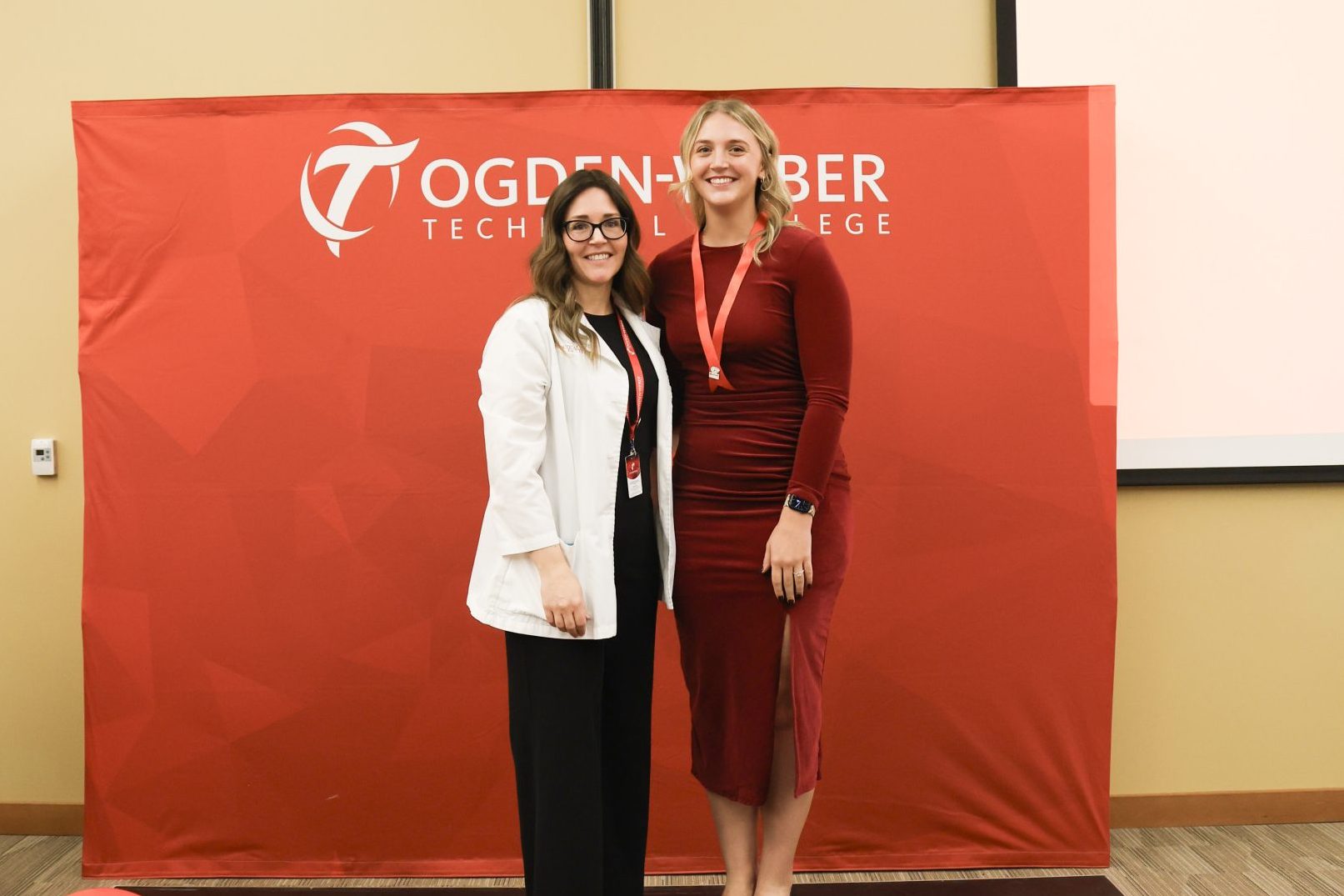 Practical nursing graduate and nursing instructor pose side in front of a red "Ogden-Weber Technical College" backdrop.