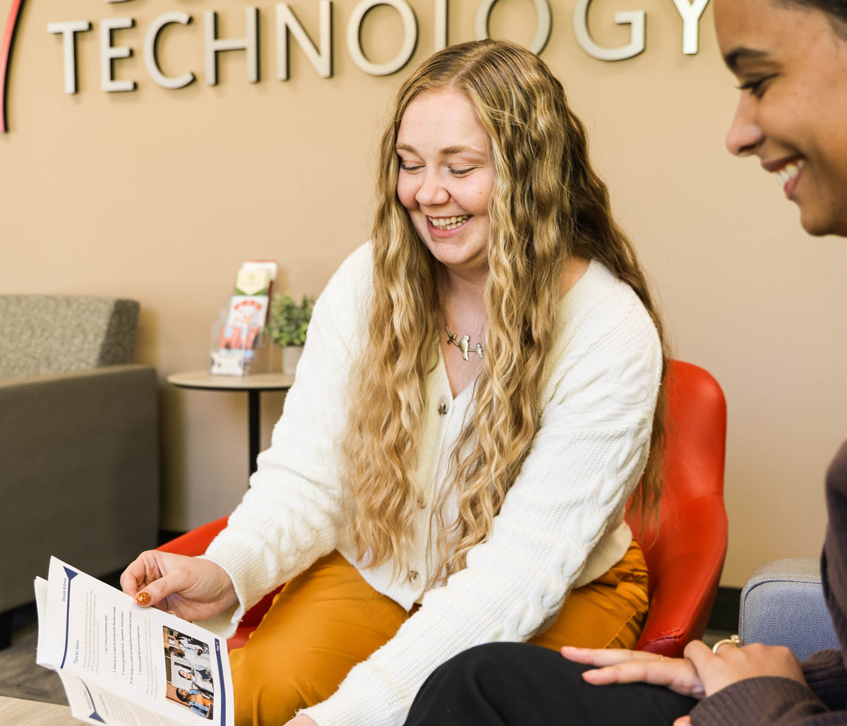 A smiling prospective student in a white sweater holds an OTECH program brochure while meeting with an admissions advisor in a bright seating area at OTECH, exploring which career training program is right for her